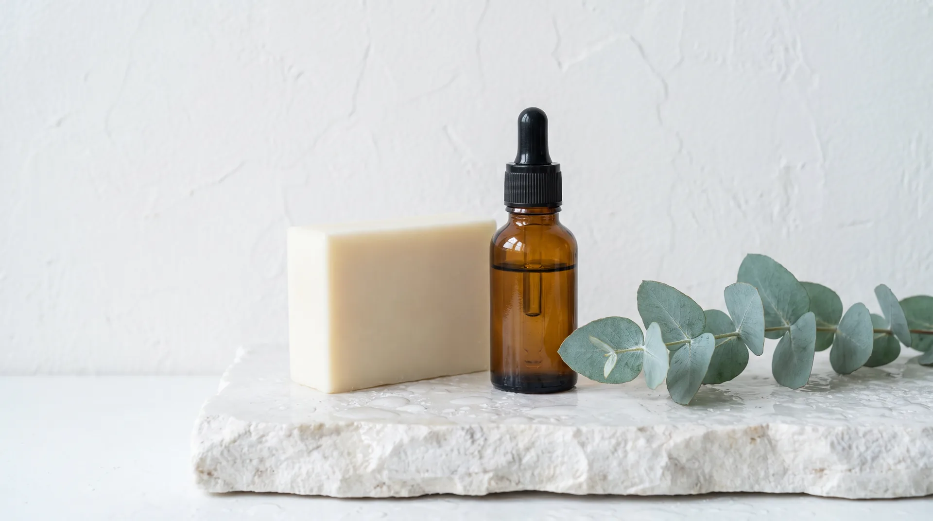 A plain cream soap bar and an amber dropper bottle on wet white stone beside a sprig of eucalyptus.