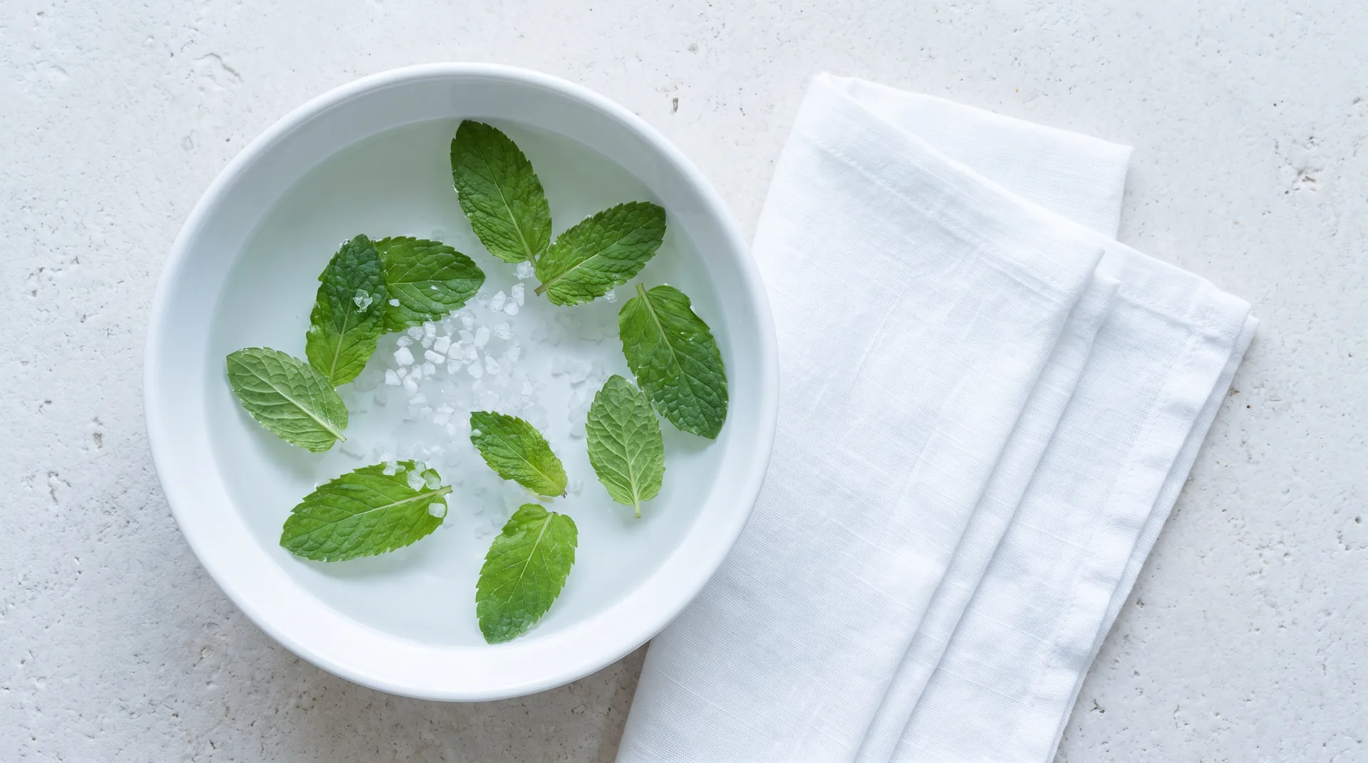 A shallow white basin of water with mint leaves and coarse salt, beside folded linen.